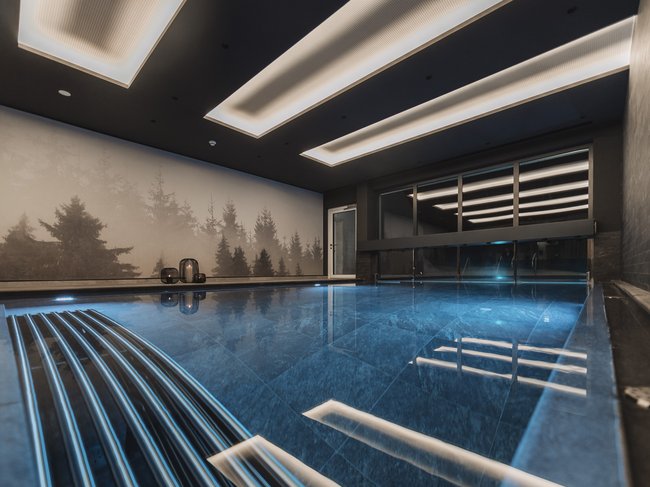 Nicht irgendein Pitztal-Resort: Alpine Resort Sportalm! Modernes Innenhallenbad mit blau beleuchtetem Wasser und Waldbild an der Wand